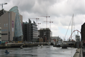 Ein Fluss mit Schiffen, eine Brücke, Gebäude, Baukräne und ein Himmel im Hintergrund.