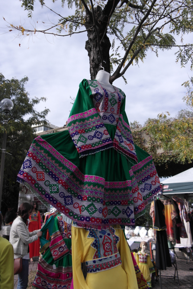 Eine Schaufensterpuppe steht in der Mitte eines Einkaufsgebiets mit mehreren Geschäften, während eine Person links Kleider an einer Stange anordnet, mit einem Gebäude und Bäumen im Hintergrund.