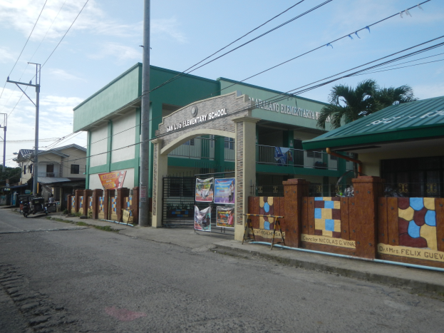 Ein Schulgebäude mit einem grünen Dach an einer Straßenecke, umgeben von Fahrzeugen, Werbetafeln, Strommasten, Kabeln, Bäumen und einem bewölkten Himmel.