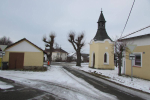 Ein kleines Dorf mit einer Kirche in der Mitte, umgeben von Häusern, Schildern, einem Mülleimer, Bäumen und Stromleitungen, alles im Schnee unter einem bewölkten Himmel.