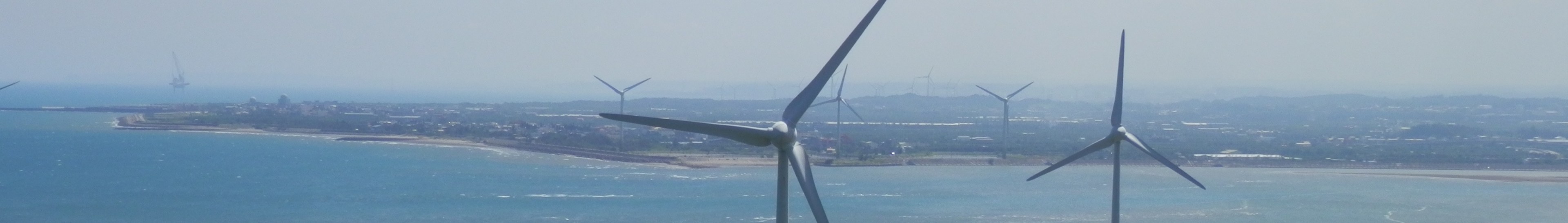 Gruppe von Windkraftanlagen im Meer in der Nähe einer Stadt, mit Wasser, das gegen die Küste schwappt, Bäumen und Gebäuden im Hintergrund und einem klaren blauen Himmel darüber.