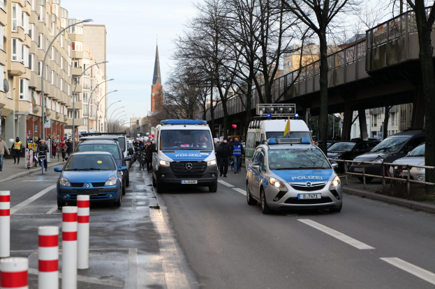 Eine Gruppe von Polizeiwagen fährt eine Straße mit hohen Gebäuden entlang, mit Menschen, die zu Fuß gehen und Fahrräder fahren, einer Brücke mit Geländern auf der rechten Seite und Bäumen mit einem klaren blauen Himmel im Hintergrund.