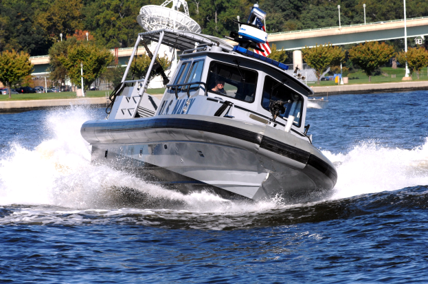 Ein Polizeiboot fährt über ein Gewässer mit einer Person darin, mit einer Brücke, Bäumen, Pfählen, Fahrzeugen, Gras und einem klaren blauen Himmel im Hintergrund.