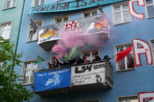 Eine Gruppe von Menschen auf einem Balkon mit Fahnen und Spruchbändern, mit einem Baum links und Rauch in der Luft, was auf eine Demonstration hindeutet.