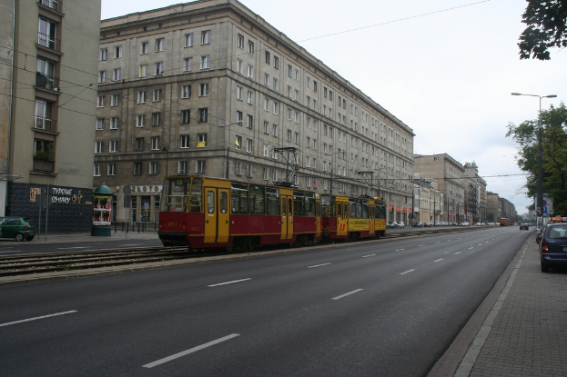 Eine Stadtansicht mit Gebäuden, Fahrzeugen, einer Bahn, Straßenlaternen, Pfählen, Kabeln, einer Straße, Bäumen, Fenstern, Geschäften und einem bewölkten Himmel.