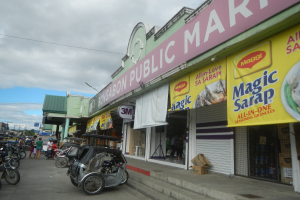 Eine belebte Straße mit parkenden Fahrzeugen, Füpßgängern auf dem Gehweg, Gebäude mit Schildern, Oberleitungen, Bäume und ein bewölktes Himmel, mit einem Laden namens "Bongabon Public Market" im Vordergrund.