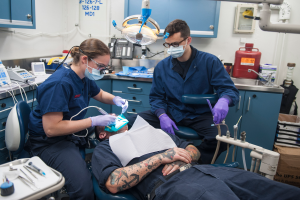 Ein Mann sitzt in einem Zahnarztstuhl, zwei maskierte und handschuhgetragene Personen versorgen ihn, eine hält ein Dentalinstrument; der Raum ist mit verschiedenen Instrumenten, Schrankwänden, einer Maschine, einem Feuerlöscher, einem Waschbecken und einer Wandtafel ausgestattet.