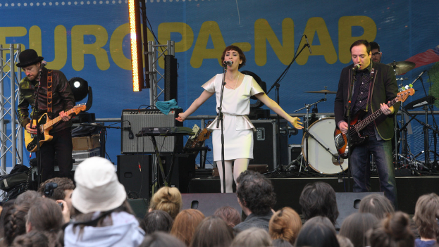 Eine Frau in einem weißen Kleid singt auf der Bühne bei einem Konzert, flankiert von zwei Gitarristen, mit einer Menge und Lautsprechern im Hintergrund.