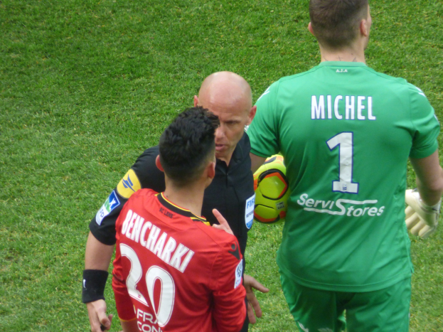 Schiedsrichter spricht mit zwei Fußballspielern auf einem Rasenfeld, ein Spieler hält einen Fußball.