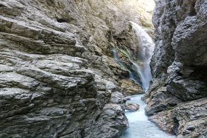 Kleiner Wasserfall, der an zerklüfteten Felsen in einem steinigen Tal hinabstürzt, umgeben von saftig grünen Hügeln, mit kristallklarem Wasser bei strahlendem Sonnenschein.