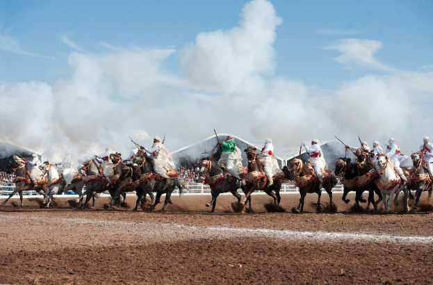 Menschen reiten auf Pferden am Boden mit anderen, Zäunen und Unterständen im Hintergrund bei bewölktem Himmel.