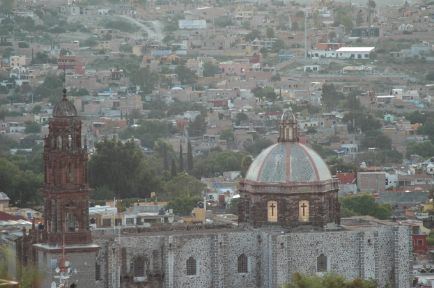 Luftaufnahme einer Stadt mit einer Kirche im Vordergrund, die Bäume, Gebäude, Straßen und Masten zeigt.