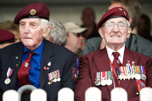Zwei Personen in formeller Kleidung mit braunen Mützen, geschmückt mit Medaillen, sitzen vor einem leicht unscharfen Hintergrund.