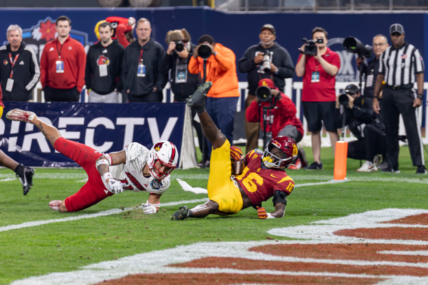 Ein Football-Spieler taucht in die Endzone für ein Touchdown, mit einer Gruppe von Menschen am Boden, einige halten Kameras, und ein Banner mit Text im Vordergrund, und Geländer, Pfosten und andere Gegenstände im Hintergrund.