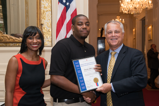 Eine Person in Schwarz und Rot lächelt links, während eine andere Person in einem schwarzen T-Shirt ein Zertifikat hält und mit einer Person in einem Anzug die Hand schüttelt, beide lachend; im Hintergrund ist eine weitere Person, Lichter auf dem Dach, ein Spiegel, eine Flagge und eine Wand zu sehen.