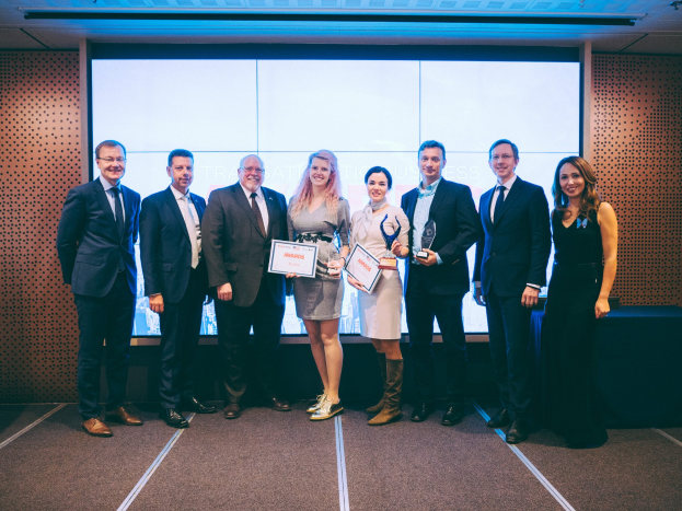 Gruppe von strahlenden Menschen mit Urkunden, die vor einem Bildschirm mit einem Tisch und einem Gegenstand im Hintergrund stehen, Gewinner der International Business Awards 2018.