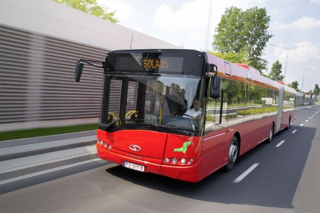 Ein roter Bus fährt auf einer Straße neben einem Gebäude, mit einer Person im Inneren, neben einer Wand, Bäumen, Laternen und einem bewölkten Himmel im Hintergrund.