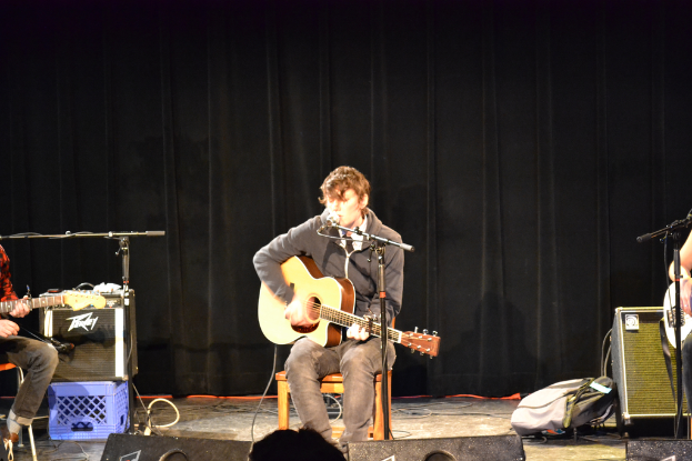 Eine Gruppe von Menschen, die vor einem Mikrofon Gitarren spielen, mit Lautsprechern, Gepäck und Vorhängen im Hintergrund.