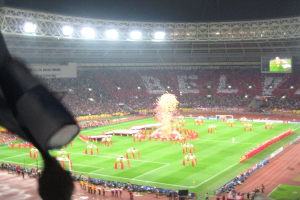 Ein Stadion mit Flutlicht, Eisengittern, Zuschauern, stehenden Personen, Werbetafeln, Anzeigetafeln, einer Überwachungskamera und verschiedenen Fahnen und Dekorationen.