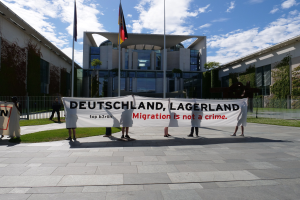 Eine Gruppe von Menschen hält eine Fahne mit der Aufschrift "Deutschland, Lagerland Migration ist kein Verbrechen" vor einem Gebäude mit Fenstern, umgeben von Gras, einem Metallzaun, Pflanzen, Bäumen und Fahnenstangen, unter einem bewölkten Himmel.