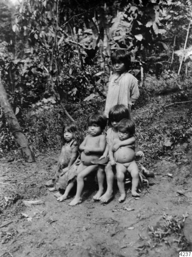 Fünf Kinder sitzen auf einer Schotterstraße in einem Schwarz-Weiß-Bild mit Bäumen und einer stehenden Person im Hintergrund und Text unten; die Kinder sehen besorgt aus.
