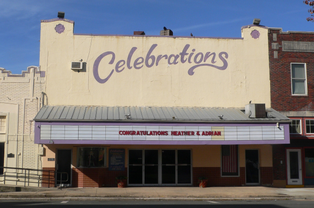 Theater mit einer "Feierlichkeiten"-Aufschrift an der Seite, umgeben von Gebäuden mit Fenstern, Türen, Geländern und Topfpflanzen, einer Straße unten, einem Baum auf der rechten Seite und einem Himmel im Hintergrund.