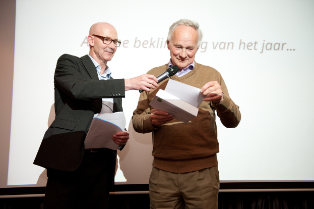 Zwei Männer stehen vor einem Bildschirm, jeder hält Papiere, wobei einer eine Brille trägt und ein Mikrofon hält.