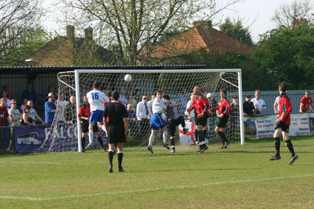 Fußballspieler sind auf einem Feld mit einem Tornetz im Einsatz, während Zuschauer dahinter stehen, mit Bäumen und Häusern im Hintergrund.