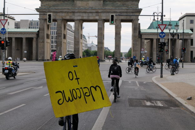 Eine Gruppe von Radfahrern mit Helmen fährt vor dem Brandenburger Tor in Berlin, Deutschland, vorbei, wobei eine Person ein gelbes Schild hält, Lichtmasten, Verkehrszeichen, Gebäude, Bäume und einen klaren blauen Himmel im Hintergrund zu sehen sind.