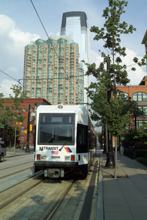 Ein Zug auf Schienen mit Stromkabeln oben, flankiert von Bäumen, Masten und Gebäuden, mit einem Wolkenkratzer und Himmel im Hintergrund.