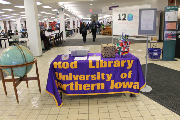Eine Ausstellung mit einem Tisch, der verschiedene Landesflaggen, eine Box, Papiere und eine Fahne zeigt, eine Kugel links daneben, eine Mitteilungstafel rechts daneben mit ein paar Menschen in der Nähe und mehrere Tische und Stühle links daneben.