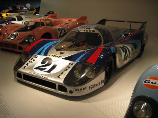 Ein Porsche 917K Le Mans Rennauto in einem Museum ausgestellt, umgeben von anderen Sportwagen, mit Menschen in der Nähe und einer Wand im Hintergrund.