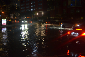 Flutstraße bei Nacht mit Autos, die durch Wasser fahren, Gebäuden, Bäumen, Laternen und Schildern im Hintergrund.