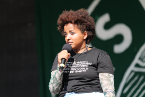 Eine Frau steht selbstbewusst auf einer Bühne, hält ein Mikrofon in der Hand und trägt ein schwarzes T-Shirt und Jeans, ihr Haar ist zu einem Pferdeschwanz gebunden.