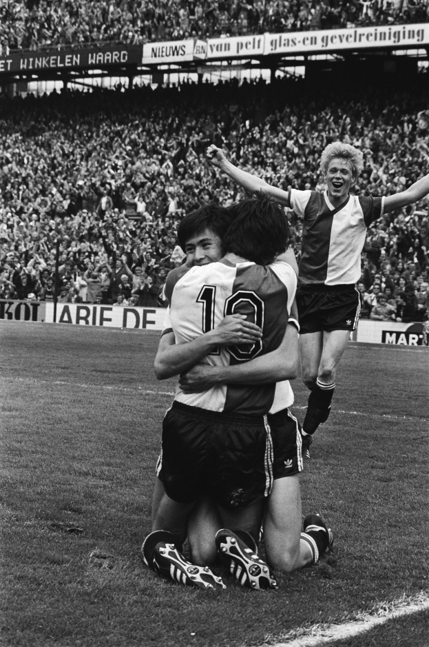 Drei Fußballspieler feiern auf dem Boden nach einem Torerfolg, mit jubelnder Menge und schwenkenden Fahnen in einer schwarz-weißen Stadionatmosphäre.