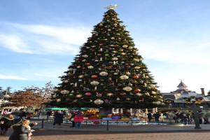 Ein dekorativer Weihnachtsbaum mit einem schwarzen Zaun davor, zwei Mädchen, die stehen und sprechen, mit einem Schloss im Hintergrund.