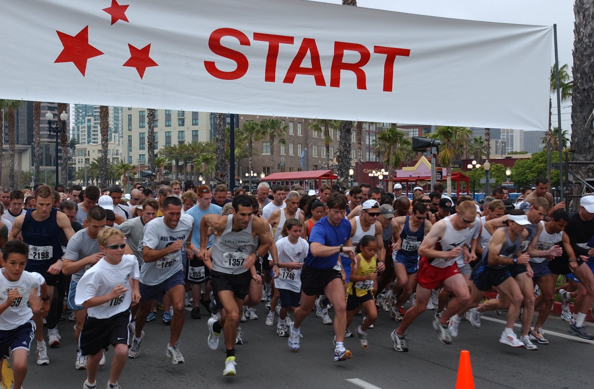 Gruppe von Menschen, die bei einem Marathon laufen, mit einem Verkehrskegel im Vordergrund und einer Fahne mit Text im Hintergrund, umgeben von Bäumen, Laternenmasten, Gebäuden und einem klaren blauen Himmel.