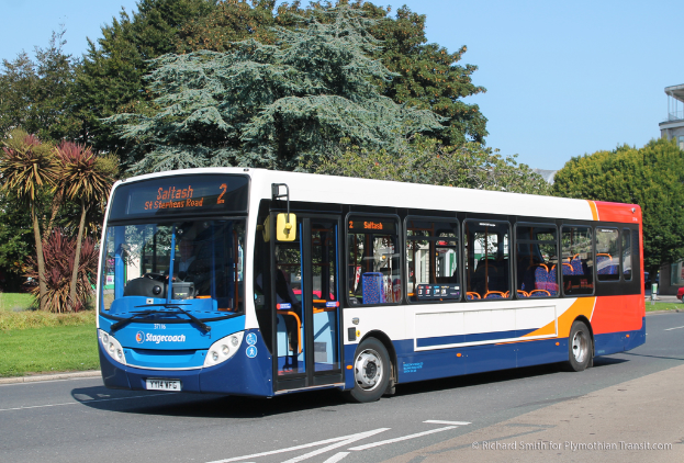 Ein Bus fahrt auf einer Stra├če mit Gras auf der rechten Seite, B├Ąumen im Hintergrund und einem Geb├Ąude auf der rechten Seite.