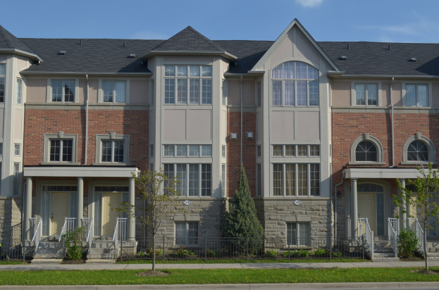 Eine Reihe von Stadthäusern in einer Wohngegend mit sichtbaren Fenstern, Türen, Säulen, Geländern, Treppen, Pflanzen, Bäumen, Gras, einer Straße im Vordergrund und dem Himmel im Hintergrund.