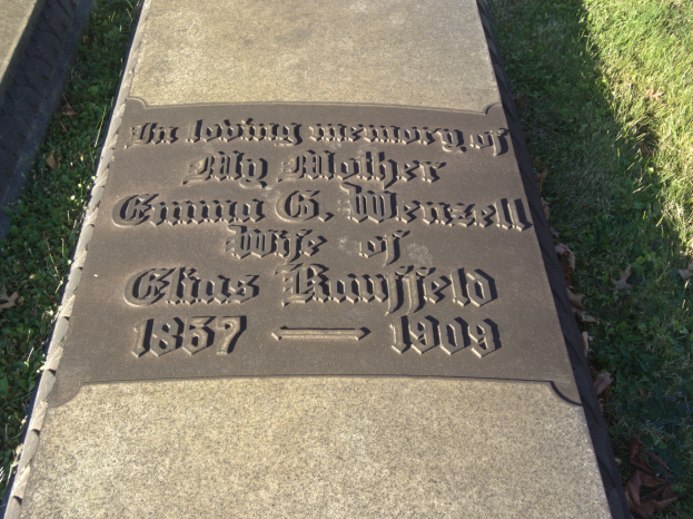 Grabstein mit der Inschrift "Die liebevolle Erinnerung an meine Mutter" in einem grasbewachsenen Friedhof mit trockenen Blättern.