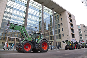 Eine Gruppe von Traktoren fährt eine Straße entlang vor einem Gebäude, mit Menschen auf dem Gehweg und einem Baum auf der rechten Seite, wahrscheinlich bei einer Demonstration.