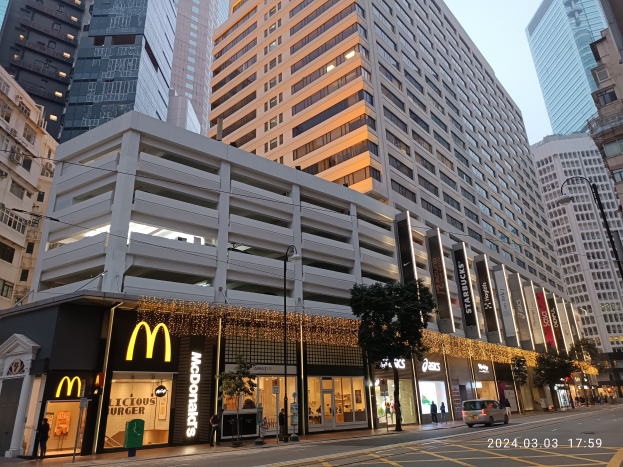 Ein McDonald's-Restaurant auf einer belebten Stadtstraße, flankiert von hohen Gebäuden, Straßenlaternen, fahrenden Fahrzeugen, Fußgängern, Bäumen, unter einem klaren blauen Himmel.