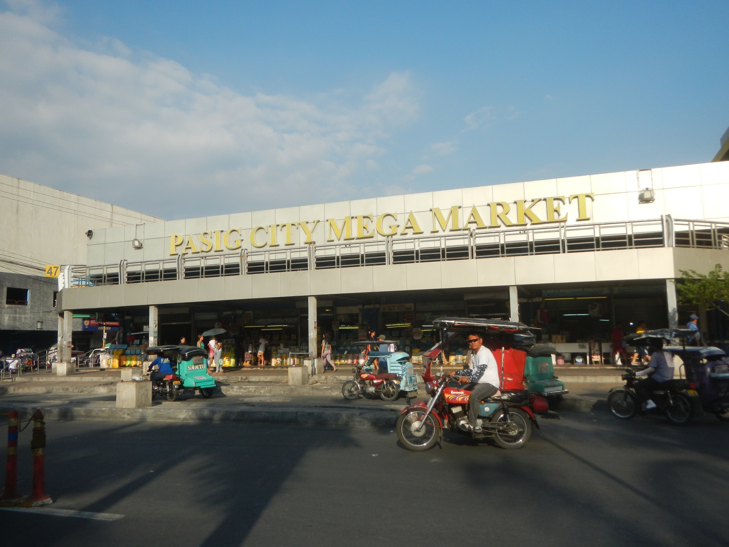 Eine belebte Straßenszene am Pasig City Mega Market mit Fahrzeugen und Fußgängern, ein Gebäude mit sichtbarem Text im Hintergrund und ein bewölkter Himmel.