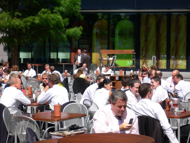 Eine Kantinenszene mit Gruppen von Menschen an Tischen sitzend und sich unterhaltend, zwei Personen in schwarzen Anzügen in der Mitte stehend, ein Flaschencontainer rechts, ein Baum links und verschiedene Gläser und Flaschen auf den Tischen.