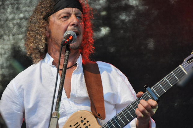 Ein Mann in einem weißen Hemd und schwarzem Kopftuch singt in ein Mikrofon und hält eine Gitarre.