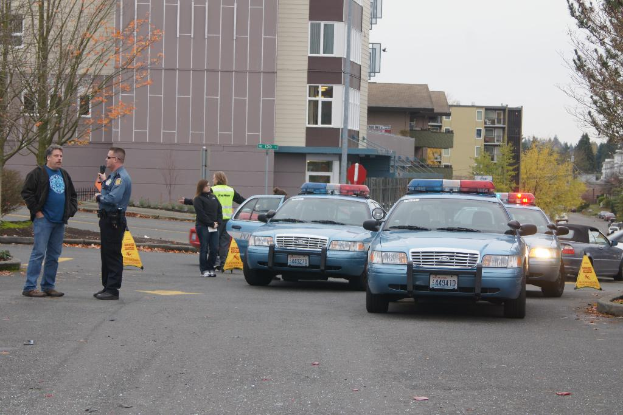 Autos auf einer Straße mit vier Personen in der Nähe, Gebäude mit Fenstern im Hintergrund, Bäume und Warndreiecke.