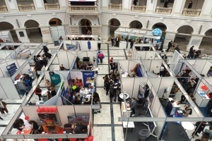 Eine gro├če Gruppe von Menschen versammelte sich in einem Raum voller St├Ąnde, Banner, Tische, St├╝hle, Luftballons und einem Geb├Ąude mit Bogen, Pfeilern, Gel├Ąndern und einem Bildschirm im Hintergrund.