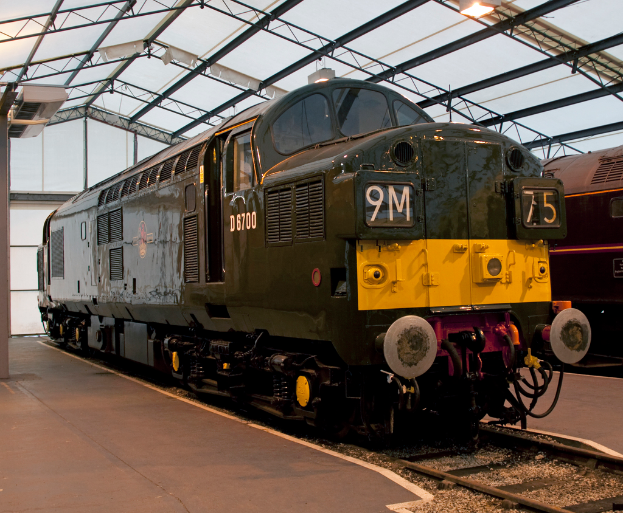 Ein Zug befindet sich auf den Gleisen neben einem Bahnsteig mit einem Mast auf dem Dach, der Metallstangen mit Lampen trägt.