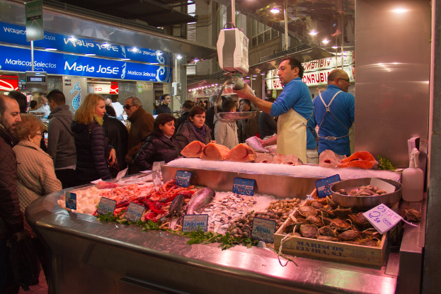 Ein Marktplatzszenario mit Menschen, die gehen und einige Fische schneiden, mit Fischen und Brettern unten und Geschäften, Brettern, Pfählen und Lichtern im Hintergrund.