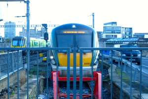 Zwei Züge auf dem Gleis mit einem Zaun im Vordergrund, Pfosten am Land, einer mit einer Signallampe, ein Auto auf der Straße rechts, Gebäude im Hintergrund und Himmel oben.